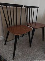 Two vintage solid wood chairs with black spindle backs and wood seats showing wear placed next to each other near a wall.