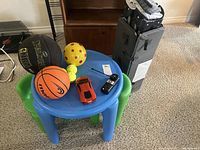 Blue Little Tikes table with assorted balls and toy cars, portable playpen nearby.