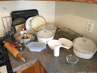 Overview of assortment on kitchen counter including Corning Ware white casseroles with lids, glass Pyrex baking dishes, metal pans, rolling pin, and small kitchen tools