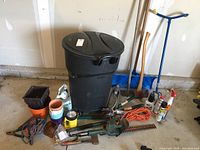 Full lot overview showing Rubbermaid trash pail, hedge trimmer, garden claw, snow shovel, flower pots, extension cord, and garden sprays.