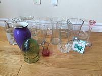 Full view of assorted glass and porcelain vases displayed on wooden floor, showing variety of shapes, colors, and sizes.