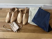 Two pairs of wooden shoetrees and two suit bags (beige and navy blue) arranged on a wooden floor.