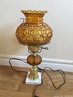 Full view of amber glass hurricane style electric lamp sitting on a white marble square base with brass metal components and attached electric cord.
