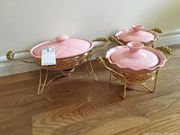 Set of three pink porcelain chafing dishes on decorative gold metal stands with candleholders underneath, placed on wooden floor.
