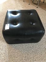 Square black faux leather ottoman showing top and front sides with button tufting and wear.