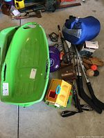 Photo showing a green plastic children's sled alongside several items including walking poles, a blue sleeping bag, binoculars, balls, and other outdoor accessories.