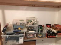 Stacked clear plastic bins containing assorted hand tools, wires, and hardware, along with gloves and a Black & Decker box.