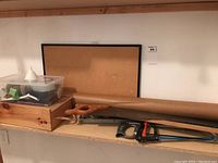 Shelf displaying wooden toolbox, clear plastic container with miscellaneous items, cork bulletin board on wall above, wood folding ruler, plastic funnel, and rolled brown paper tube.