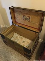 Open wooden chest showing interior with printed scenic label on lid and floral fabric lining on bottom, along with visible water damage.