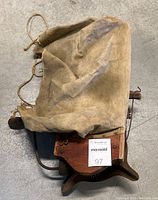 Wooden hand crank seed spreader with a canvas bag and shoulder rope laid flat on the floor, showing the crank handle and bag attachment.