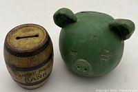 Photo showing the Happy Days Tin Bank barrel shaped bank alongside the green ceramic piggy bank, illustrating bank styles and size comparison.
