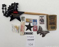 Photo of assorted small vintage advertisements, pins including a silver-tone butterfly, a black star pin, a pack of Sweet Caporal cigars, and a Black Budweiser Select star pendant necklace with black beads.