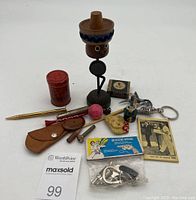Photo showing multiple small knick knacks including wooden figurine with sombrero, gold pen, red container, pins, and keychains on white background.