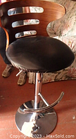 Close-up of single bar stool showing wooden backrest with three horizontal cut-outs and black padded seat with chrome base and footrest.