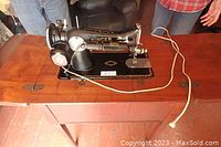 Front view of the antique black Singer electric sewing machine on the wooden cabinet showing the machine and the long power cord.
