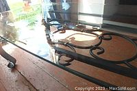 Side angle view of clear rectangular glass top showing ornamental wrought iron design underneath and portion of floor.