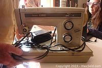 Side view of beige-colored Sears Kenmore sewing machine showing brand name and front control dials with attached power cord and foot pedal cord.