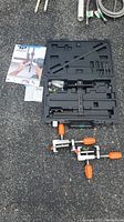 Overview of open plastic case showing drilling jig kit and two clamps on asphalt surface