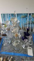 Full lot showing five glass decanters, two clear glass candlesticks, two blue glass candlesticks on a table with a forest background painting.