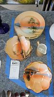 All dishes including small bowls, plates, and creamer displayed on a patio table; scenic hand-painted designs visible on most items; creamer visible as distinct piece