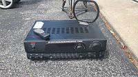 Full view of the black Pyle PT260A stereo receiver with remote control placed on top, showing vehicle pavement background.