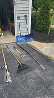 Full view of seven yard tools laid on driveway in front of garage, including two snow shovels and tree pruner