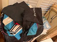 Photo showing a pile of fabric bags including several black Pandora branded tote bags and a red purse, with linens stacked nearby.