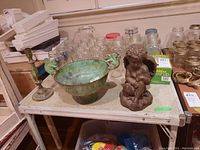Photo of metal candlestick, metal bowl with bird handles, and angel doorstop figurine on a table surrounded by jars and other items