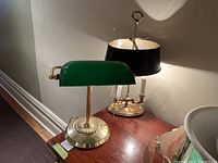 Photo showing both lamps on a wooden surface, the front lamp with green glass shade and polished brass base and stem, the back lamp with black metal shade and brass base with candle-like bulb holders.