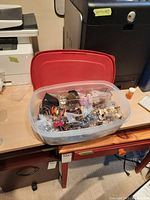 Plastic storage container holding multiple pieces of assorted costume jewelry including bracelets, necklaces, rings, and a key chain with a red lid beside it.