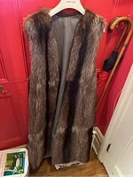Front view of the sleeveless raccoon fur vest on a hanger against a red door, showing the full length and the fur texture along with suede ties and silk lining at the edges.