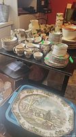Full display of over 50 pieces of Johnson Bros Ironstone Friendly Village dinnerware, including stacked plates, cups, creamers, and sugar bowls on a black table.