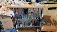 Front and side angle view of butcher block top kitchen cart with metal frame and wire shelves on caster wheels, displaying glass bottles on shelves and tea cups on top.