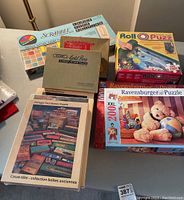 Lot of puzzles on a table including Ravensburger 200 piece XXL puzzle featuring teddy bears, vintage tool box puzzles, Lee Valley unopened and opened puzzles, RollOPuzz mat, and Scrabble game box.