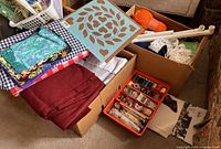 Wide view showing boxes containing assorted quilting fabrics, plastic frame, blue quilting template, orange yarn, and sewing notions kit