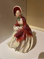 Front angle showing seated woman figurine dressed in bonnet, red shawl, and white dress with yellow accents.