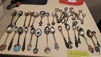 Overview of 25 souvenir spoons laid out on a white surface showing varied designs and handle decorations.