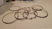 Eight sterling silver bangles arranged loosely on a flat surface. Various designs visible including plain polished and patterned. Some have clasps and hinges.