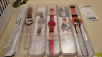 Lot of 5 Swatch watches each in individual clear plastic cases, arranged side by side showing all watches and some certificates beneath.