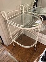 White painted metal tea cart with two clear glass shelves, decorative metal scrollwork, and metal caster wheels, shown against hardwood flooring and mirrored wall.