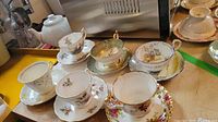 Photo showing six different fine china teacups and saucers with various floral patterns and gold trim arranged on a wooden surface