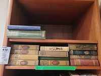 Wide shot of 14 vintage hardcover books on a wooden shelf. 10 books are laid horizontally on one shelf, 4 vertically on another. Books vary in condition and cover colors including beige, green, and blue.