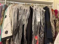 Collection of women's jackets and tops on hangers, some protected in plastic garment bags, showcasing variety of colors and fabrics.