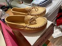 Pair of light tan leather women's moccasins displayed side by side on top of their original box, showing the whipstitched front and tied lace details.