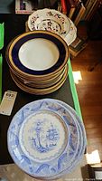 Stack of mixed porcelain plates including floral, blue rim, and blue and white Chinese design plates.