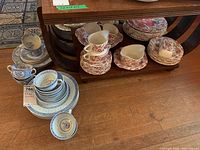 Photo showing two different china patterns on a wooden floor and shelf, with blue and white plates, cups, and saucers stacked on the floor and red patterned set on the shelf.