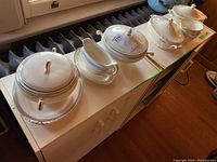 Lidded serving bowls, gravy boat with plate, and soup bowls on cabinet near a radiator, showing complete set.