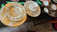Three sets of fine bone china arranged: Royal Crown Derby Aves Gold plates left, Caverswall Gold Geisha plates and teacup middle, Caverswall Black Imperial plates and cups right