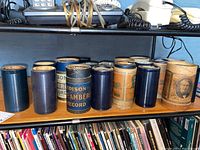 Photo showing a row of vintage wax cylinder phonograph records in their cylindrical paper containers, labeled Edison Amberol and others.