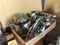 Box containing various stainless steel saucepans with lids and a strainer piled inside.